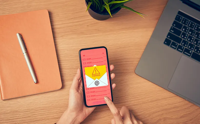 Eine Hand hält ein Smartphone, auf dem eine Malware-Warnung in einer E-Mail angezeigt wird. Auf dem Tisch liegen ein Laptop, ein Notizbuch und ein Stift.