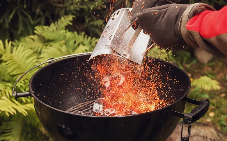  Person, die heiße Kohlen aus einem Anzündkamin in einen runden Grill schüttet. 