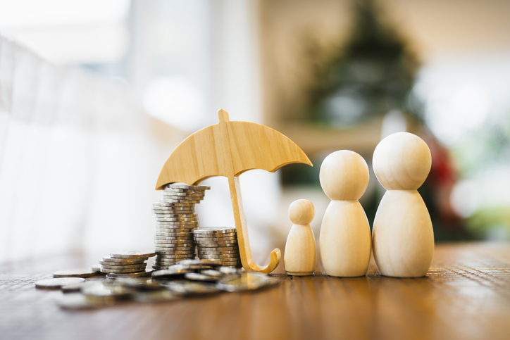 Holzfiguren einer Familie neben Geldstapeln und einem schützenden Schirm als Symbol für finanzielle Sicherheit und Versicherung