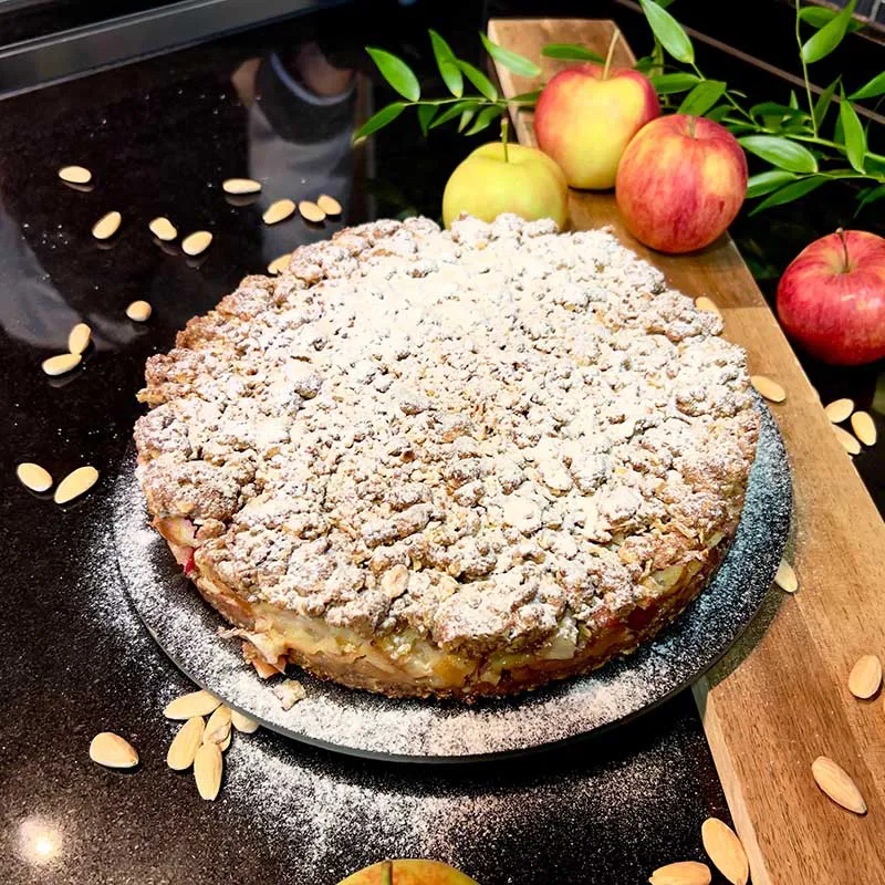 Ein Apfel-Crumble mit knusprigen Streuseln und Puderzucker, serviert auf einer schwarzen Platte, daneben frische Äpfel und Mandelblättchen zur Dekoration.
