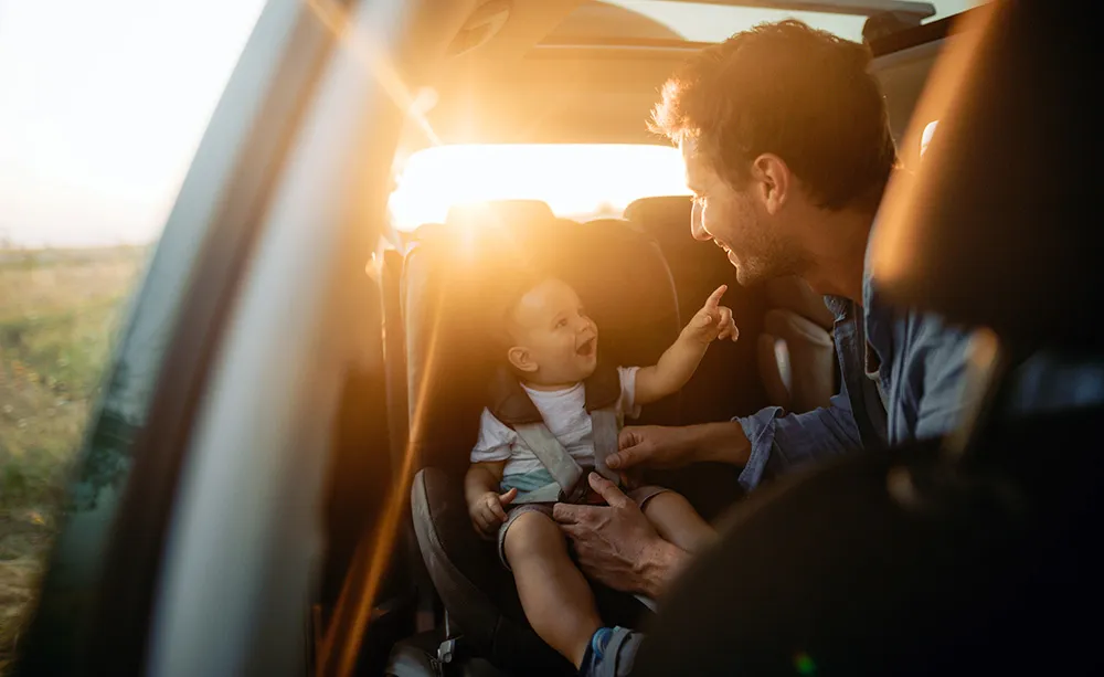 Vater interagiert lächelnd mit seinem Kind, das in einem Autositz angeschnallt ist, während die Sonne durch das Autofenster scheint