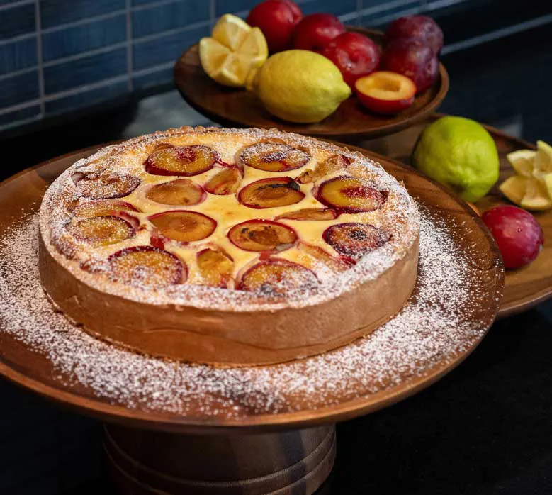 Pflaumen-Quarkkuchen, dekoriert mit Puderzucker, serviert auf einem Holzständer. Im Hintergrund liegen frische Zitronen und Pflaumen.