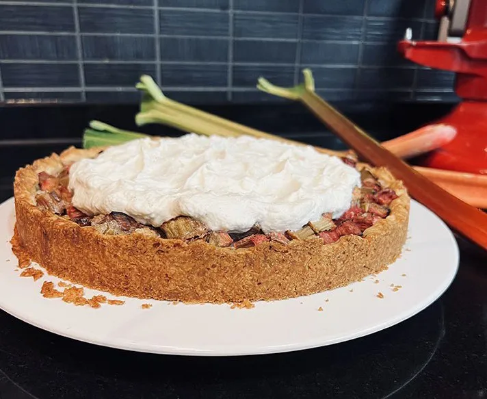 Rhabarberkuchen mit knusprigem Boden und Sahnehaube auf einem weißen Teller.