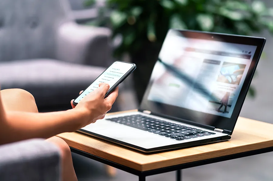 Person hat ein Smartphone in der Hand und sitzt vor einem Laptop. 