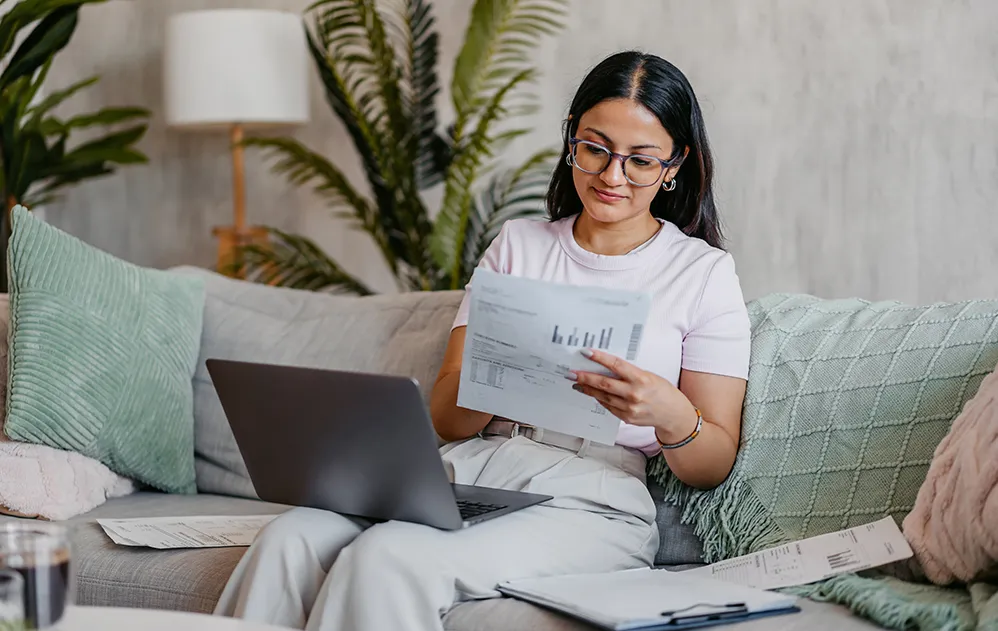 Eine Frau sitzt auf einem Sofa, einen Laptop und mehrere Bögen Papier vor sich.
