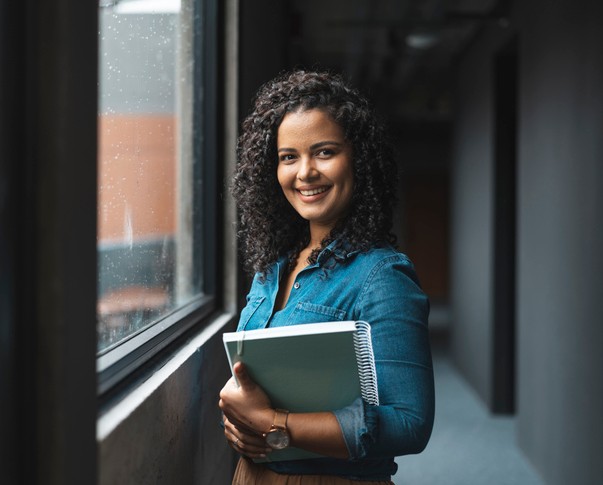 Eine junge Frau mit einem Schreibblock in der Hand steht in einem Büroflur und lächelt.