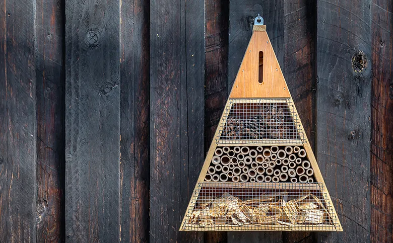 Dreieckiges Insektenhotel aus Holz, aufgehängt an einer dunklen Holzwand.