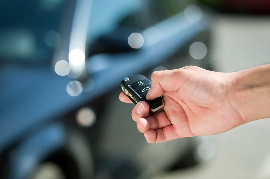 Im Vordergrund sieht man eine Hand mit Autoschlüssel, im Hintergrund steht ein Auto.