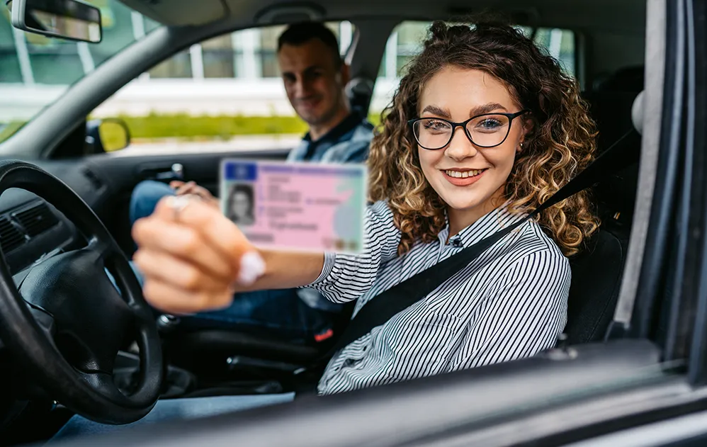 Eine junge Frau sitzt im Auto und hält ihren Führerschein in die Kamera.