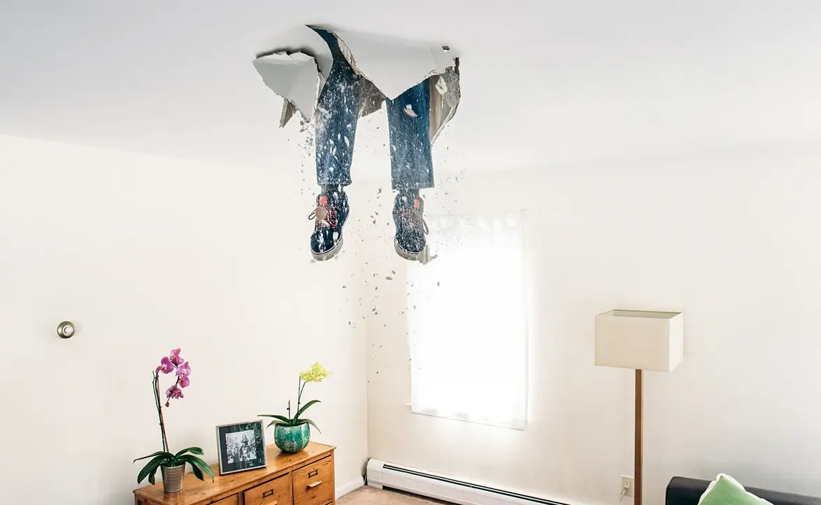 Beine einer Person ragen durch ein Loch in der Zimmerdecke, Staub und Trümmer fallen in ein helles Wohnzimmer.