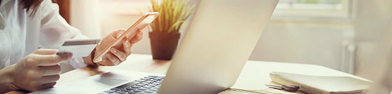 Person sitzt am Schreibtisch mit Laptop, hält Kreditkarte in der einen Hand und Smartphone in der anderen.