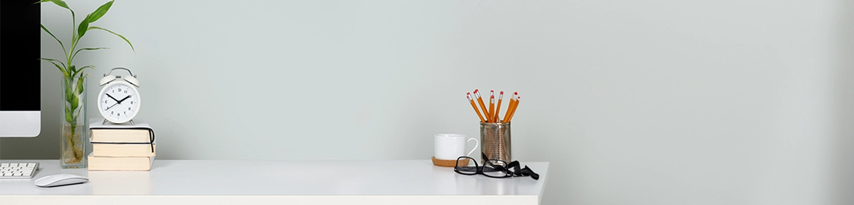 Aufgeräumter Schreibtisch mit Computer, Büchern, Stiftehalter, Brille, Uhr und Pflanze