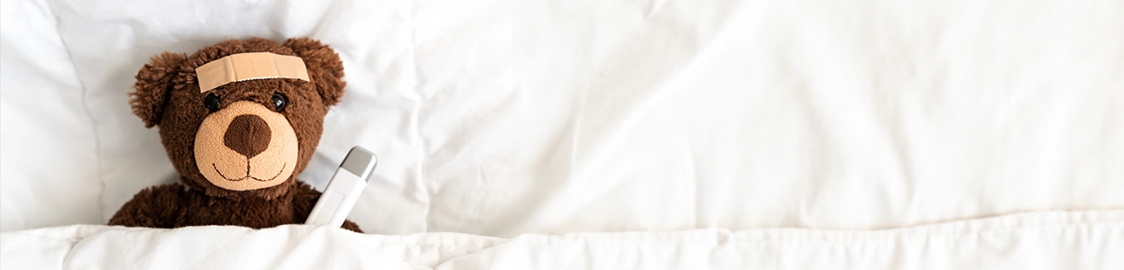 Plüschteddy mit Pflaster auf der Stirn und Thermometer im Bett liegend