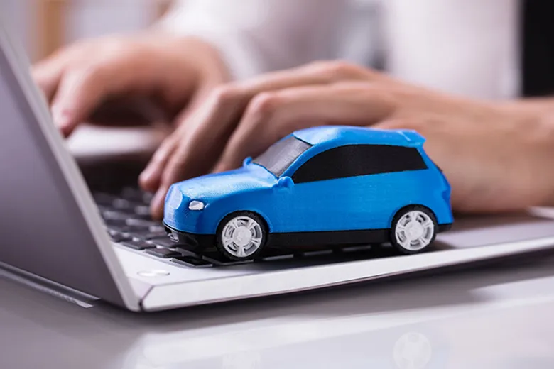 Blaues Spielzeugauto steht auf einer Laptop-Tastatur, im Hintergrund tippt eine Person