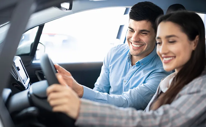 Junges Paar sitzt gemeinsam im Auto, beide lächeln und die Frau hält das Lenkrad