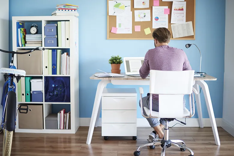Mann arbeitet im Homeoffice an einem Schreibtisch mit Laptop, Pinnwand und Regal voller Ordner