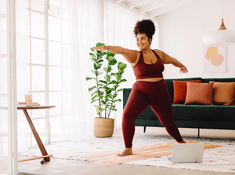 Frau in Sportkleidung macht Yogaübung im Wohnzimmer auf einer Matte, vor ihr steht ein Laptop
