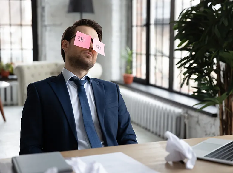 Mann im Anzug schläft am Schreibtisch, auf seinen Augen kleben Post-its mit gemalten Augen