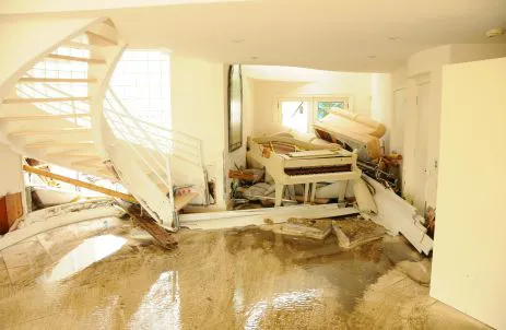 Überschwemmtes Wohnzimmer mit eingestürzter Treppe, beschädigten Möbeln und Flügel nach Hochwasserschaden