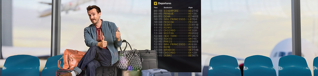Ein freundlicher Mann sitzt auf seinen Koffern am Flughafen und zeigt zwei Daumenhoch.