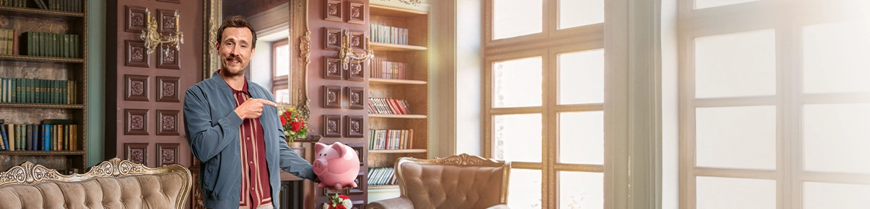 Ein lächelnder Mann steht in einem schönen Wohnzimmer und ein ein gefülltes Sparschwein in der Hand.