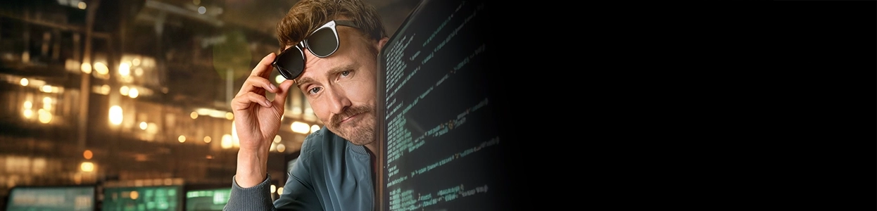 Ein freundlicher Mann schaut lächelnd hinter einem Computerbildschirm hervor und zieht seine Sonnenbrille hoch.