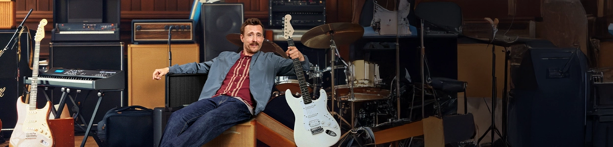 Ein lächelnder Mann sitzt zwischen einer Vielzahl von Musikinstrumenten.