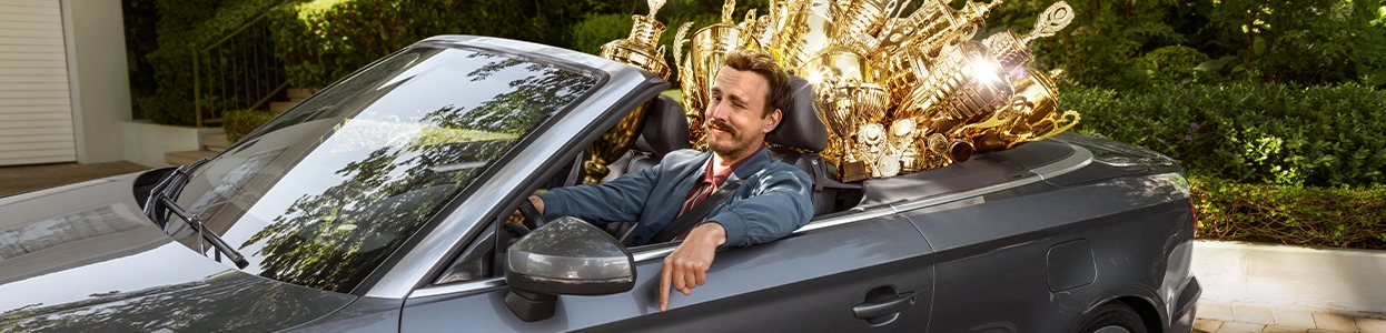 Mann fährt entspannt ein Cabrio, auf dem Rücksitz stapeln sich glänzende goldene Pokale