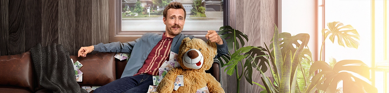 Ein entspannter Mann sitzt mit einem Teddybären voller Geldscheine auf einem Sofa.