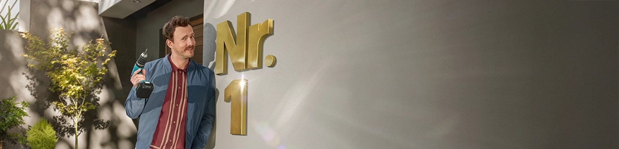 Ein lächelnder Mann mit einem Werkzeug in der Hand steht vor der goldenen Hausnummer 1.