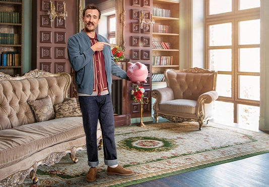 Ein lächelnder Mann steht in einem schönen Wohnzimmer und ein ein gefülltes Sparschwein in der Hand.
