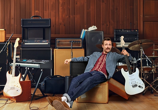 Ein lächelnder Mann sitzt zwischen einer Vielzahl von Musikinstrumenten.