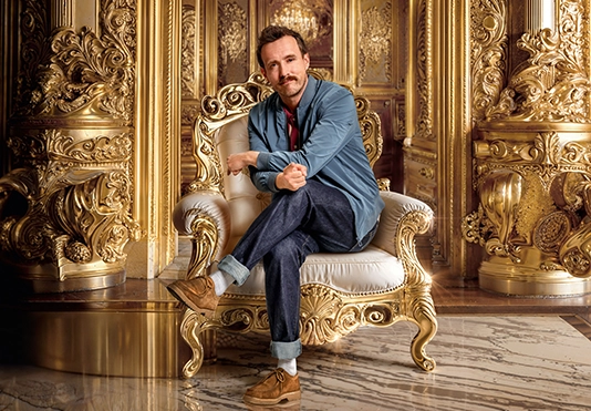 Ein lächelnder Mann sitzt auf einem goldenen Stuhl in einem prunkvollen Raum.