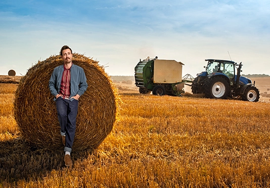 Ein lächelnder Mann steht auf einem Feld vor einem Heuball, ein Sonderfahrzeug im Hintergrund.
