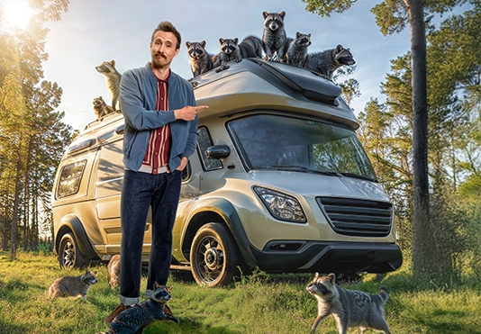 Ein freundlicher Mann steht vor einem Wohnmobil, auf dem eine große Waschbärfamilie sitzt.