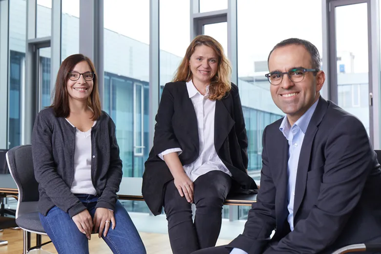 Zwei Frauen und ein Mann in Businesskleidung sitzen und lächeln in einem modernen Büro mit großen Fenstern